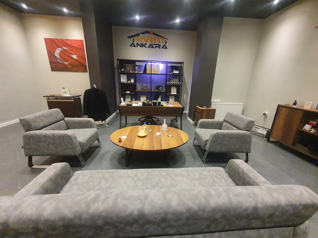 Evim Ankara ofisinin içi: gri koltuklar, yuvarlak ahşap sehpa, arkada duvarda logo